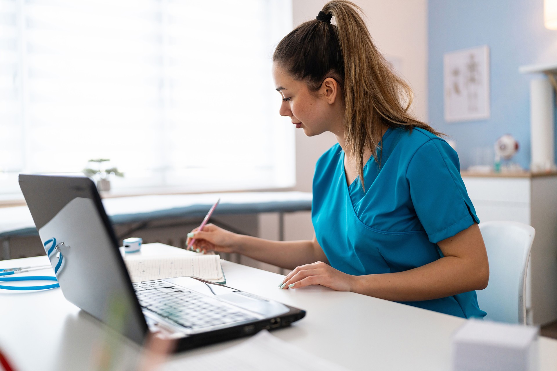 Female medical intern in blue medical scrubs e-learning on laptop at the ordination